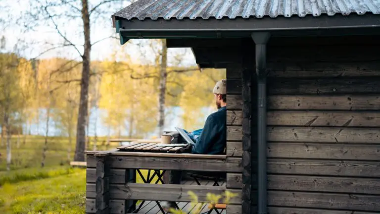 man working on laptop on a cabin porch by a lake
